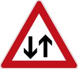 Verkehrszeichen 125 „Gegenverkehr“ mit weißem Hintergrund und rotem Rand, das zwei schwarze Pfeile zeigt, einer nach oben und einer nach unten.