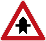 Verkehrszeichen 301 „Vorfahrt“: dreieckig mit rotem Rand und weißem Grund, das schwarze Sinnbild stellt eine Straßenkreuzung dar, wobei ein senkrechter Pfeil (Vorfahrtstraße) von einem waagerechten Strich (kreuzende oder einmündende Straße) gekreuzt wird.