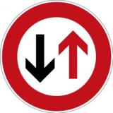 Verkehrszeichen 208 „Vorrang des Gegenverkehrs“: ein roter nach oben und ein schwarzer nach unten gerichteten Pfeil, in einem weißen Kreis mit rotem Rand.
